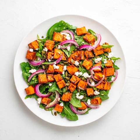 Golden roasted sweet potato salad with creamy feta, peppery arugula, and honey-lime dressing, served in a white bowl.